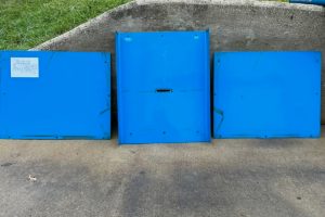 1a - Dupont Welder Services Three blue metal panels leaning against a concrete curb, left panel has a white label, on a pavement with grass behind.