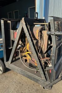Open-frame electrical transformer or coil assembly on a wheeled cart outdoors, with many yellow and colored wires.