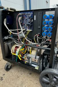 Open electrical cabinet on a wheeled cart, showing a circuit board, transformers, and a tangle of wires and connectors.