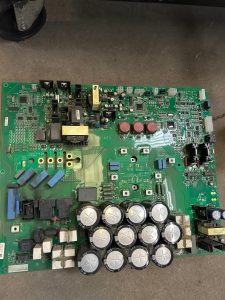 Green electronic circuit board populated with capacitors, transformers, and connectors on a concrete floor.