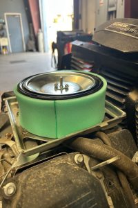 Green cylindrical air filter sits atop a small engine, with a Kohler Command Pro label visible nearby in a workshop setting.