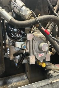 Close-up of a gas pressure regulator connected to metal piping with hoses and wires in an equipment compartment.